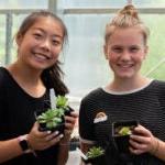 两名高中生在温室里拿着小植物微笑着拍照.